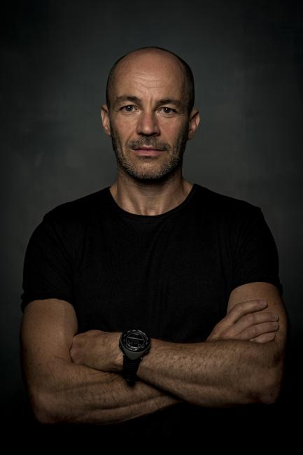 a bald man standing against a dark background with his arms crossed. He is wearing a fitted black T-shirt and a dark writswatch. 