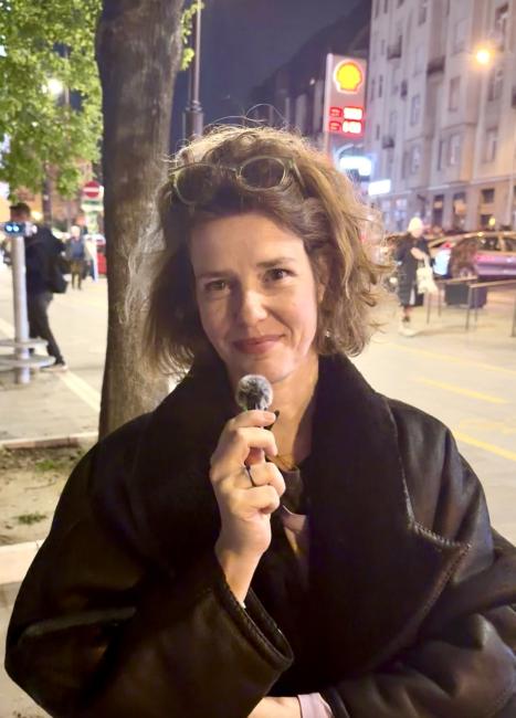 A woman with a shoulder long braing hair and sunglasses in her hair smiles and holds a small mic in her hands. She wears a black coat. 
