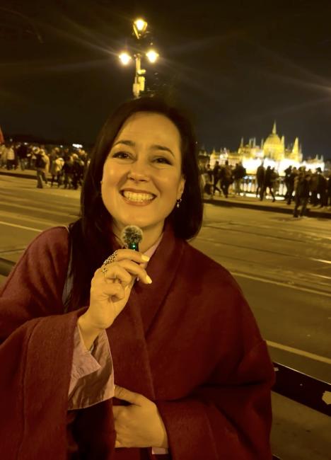A smiling young woman in a pink coat and long black hair speaking into a microphon in front of the Houses of the Parliament in Budapest. 