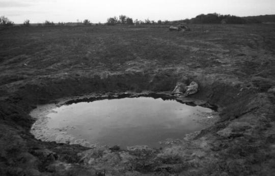 a large muddy crater filled with still water in an open field. The scene feels desolate. 