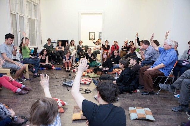 A large group of people sit in a wide circle suggesting a group session. There is a vote, some people raise their hand. The sphere is communal. 
