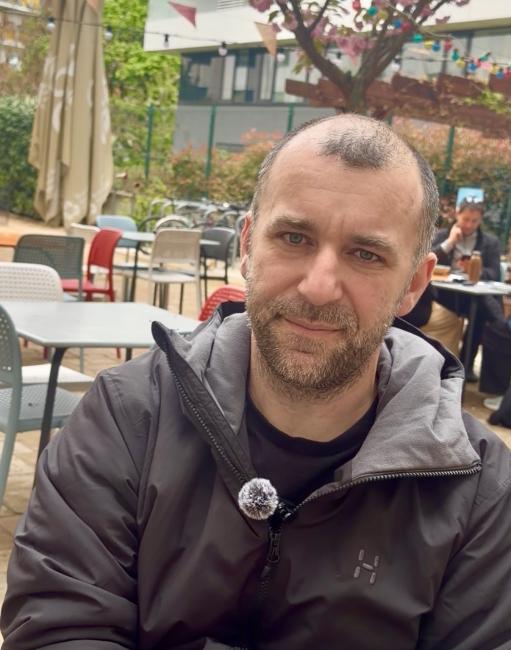 A bold man in a grey coat sitting in a garden pub, slightly smiling and looking into the camera