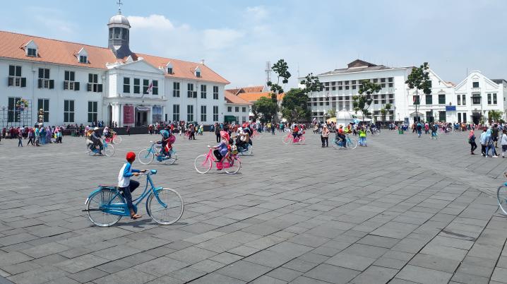 A vast square with many people, some of them on brightly coloured bikes, and buildings in the background