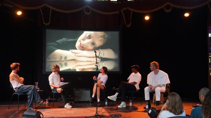 A group of 5 young people sitting on a stage watching images on a large screen
