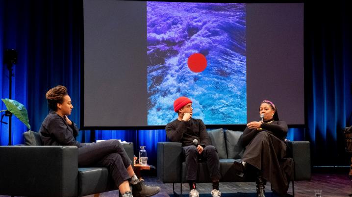 Three people as part of a panel sitting on stage. Behind them is a screen projecting an image of waves with a red dot in the middle