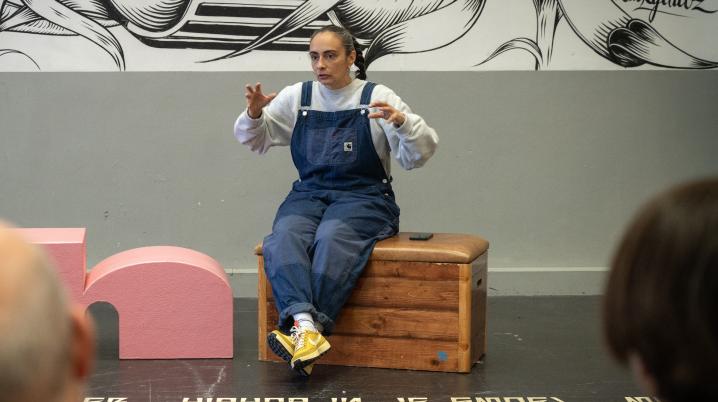 A woman sitting in a room on a wooden stool, gesturing to an audience, with a black and white grafitti art work in the background, and white lyrics written on the black floor