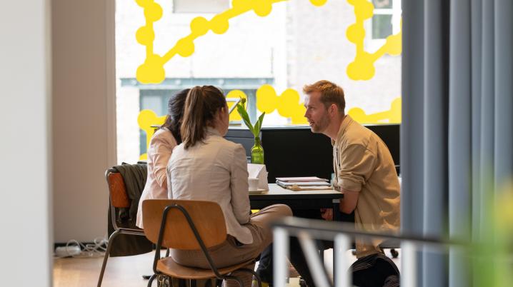 Three people in conversation sitting around a table on brown chairs. natural lighting comes in through a large window that is decorated with yellow dots