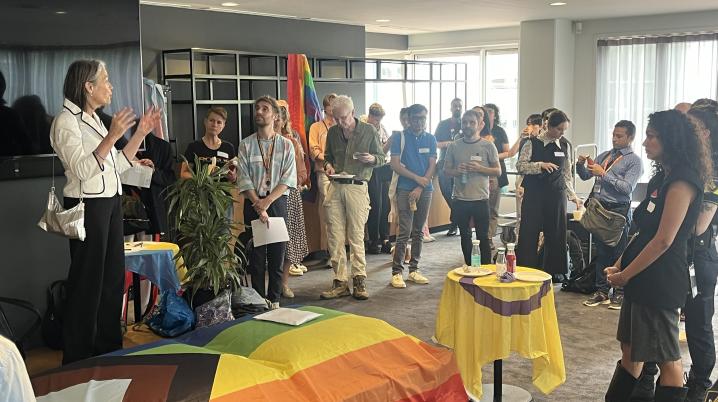 A standing group of people listen to a woman speaking, surrounded by several rainbow flags