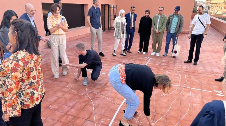 A group of people standing on a rooftop looking at a woman making a chalk drawing on the floortiles