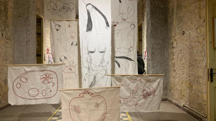 Multiple sheets of white white textile embroidered with images of pomegranates and women. The textiles are suspended from the ceiling of a galley space of which the walls are bare bricks and the floor is made of black and white tiles.