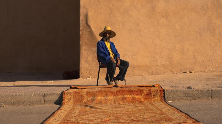 Person on a chair the side of the pavement selling a carpet