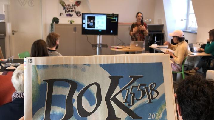 Person holding a presentation in from of people sitting down in an office. Photografer holding the newspaper of the cultural organisation DoK15518 in front of the camera.