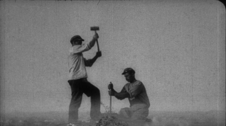 Black-and-white film still: two workers hammer a large stake atop a mound, shot in forced perspective so they appear giant over a landscape below. 