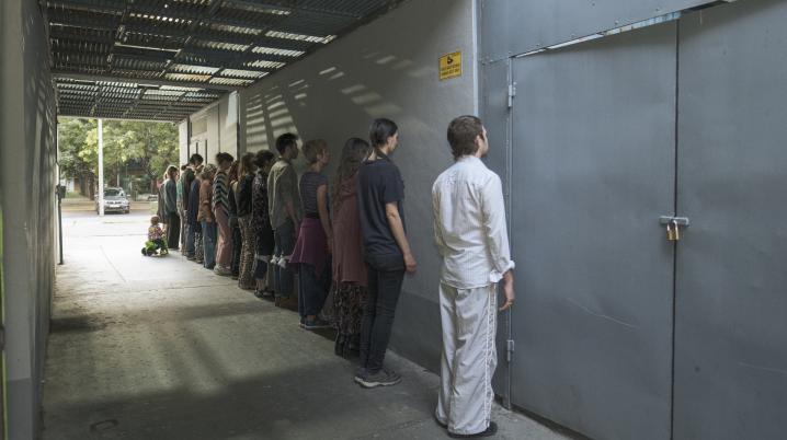 A group of people stand in a line facing a gray wall in a covered outdoor corridor. 